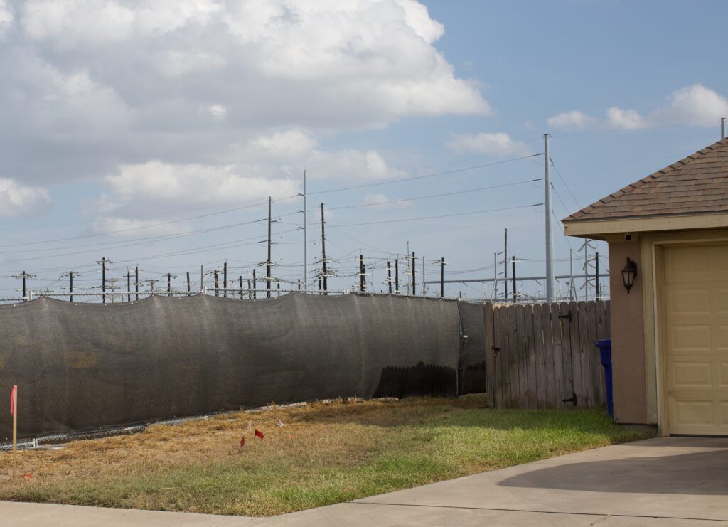 There's a single family home to the right of an industrial electric substation behind a fence on Buddy Owens Boulevard in McAllen, Texas owned and operated by AEP Texas with a blue sky in the background on a cloudy day.