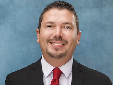 Portrait of Ediel Barrera, director on the Rio Grande City Economic Development Corporation, wearing a black suit, white shirt, and red tie.