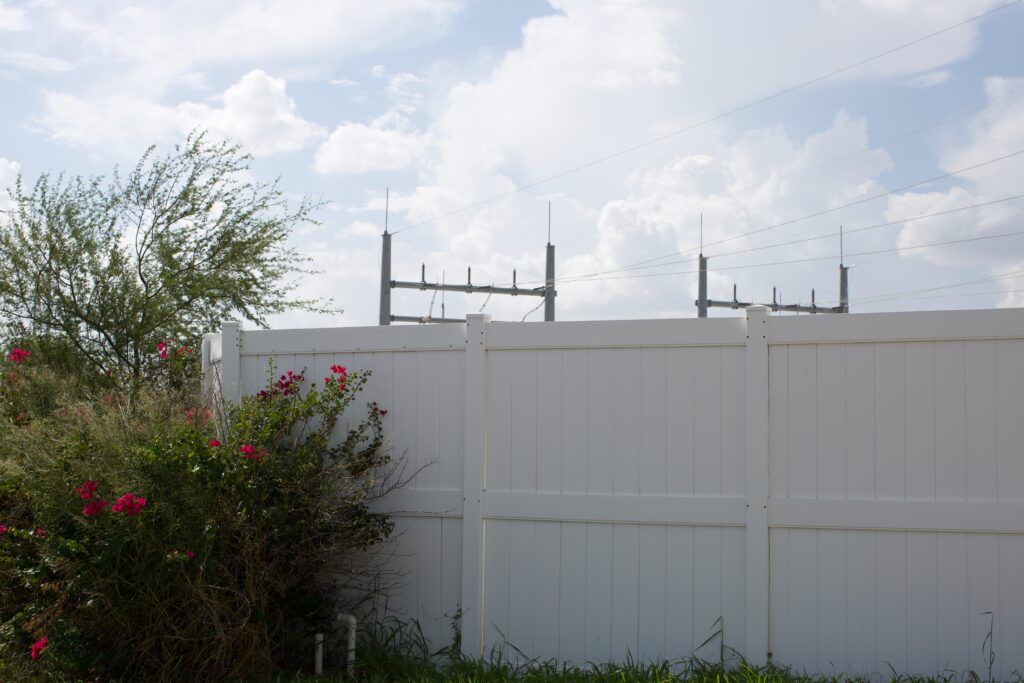 Flowering shrubs are planted next to a white picket fence that partially hides an electric substation near the border of McAllen and Pharr against a cloudy blue sky.