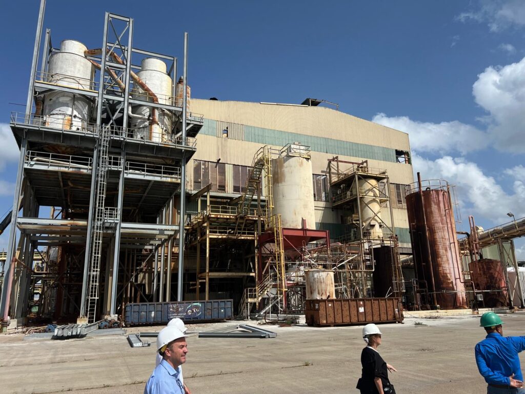 Exterior of the shuttered Santa Rosa sugar mill with workers in hard hats walking near large metal tanks and rusted structures.