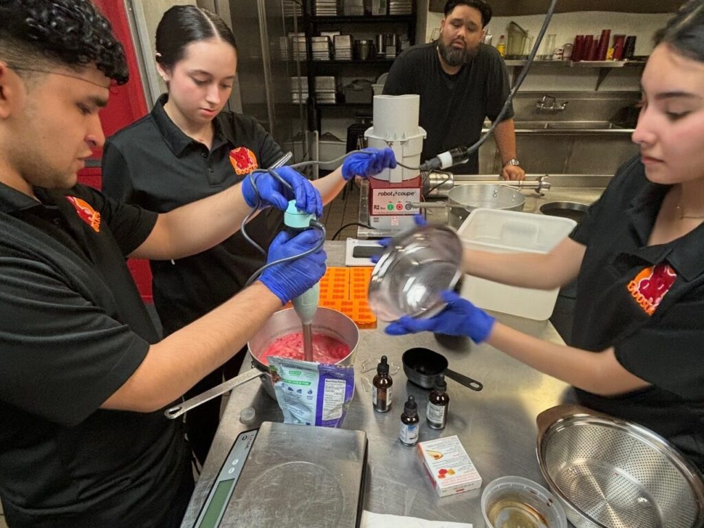 Three students are mixing red gelatin in a commercial kitchen as an adult looks on.