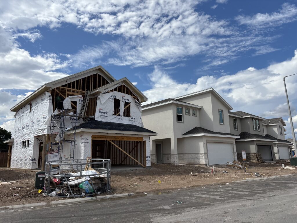 A row of two-story houses under construction at Villas on Ware in McAllen, with one home partially framed and wrapped in Tyvek material under a bright blue sky.