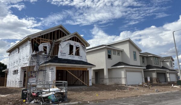 A row of two-story houses under construction at Villas on Ware in McAllen, with one home partially framed and wrapped in Tyvek material under a bright blue sky.
