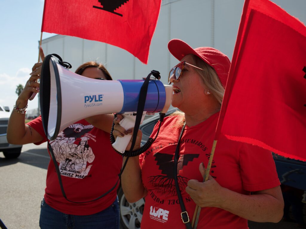 A woman wearing a red hat, a red shirt and holding a red flag shouts into a megaphone.