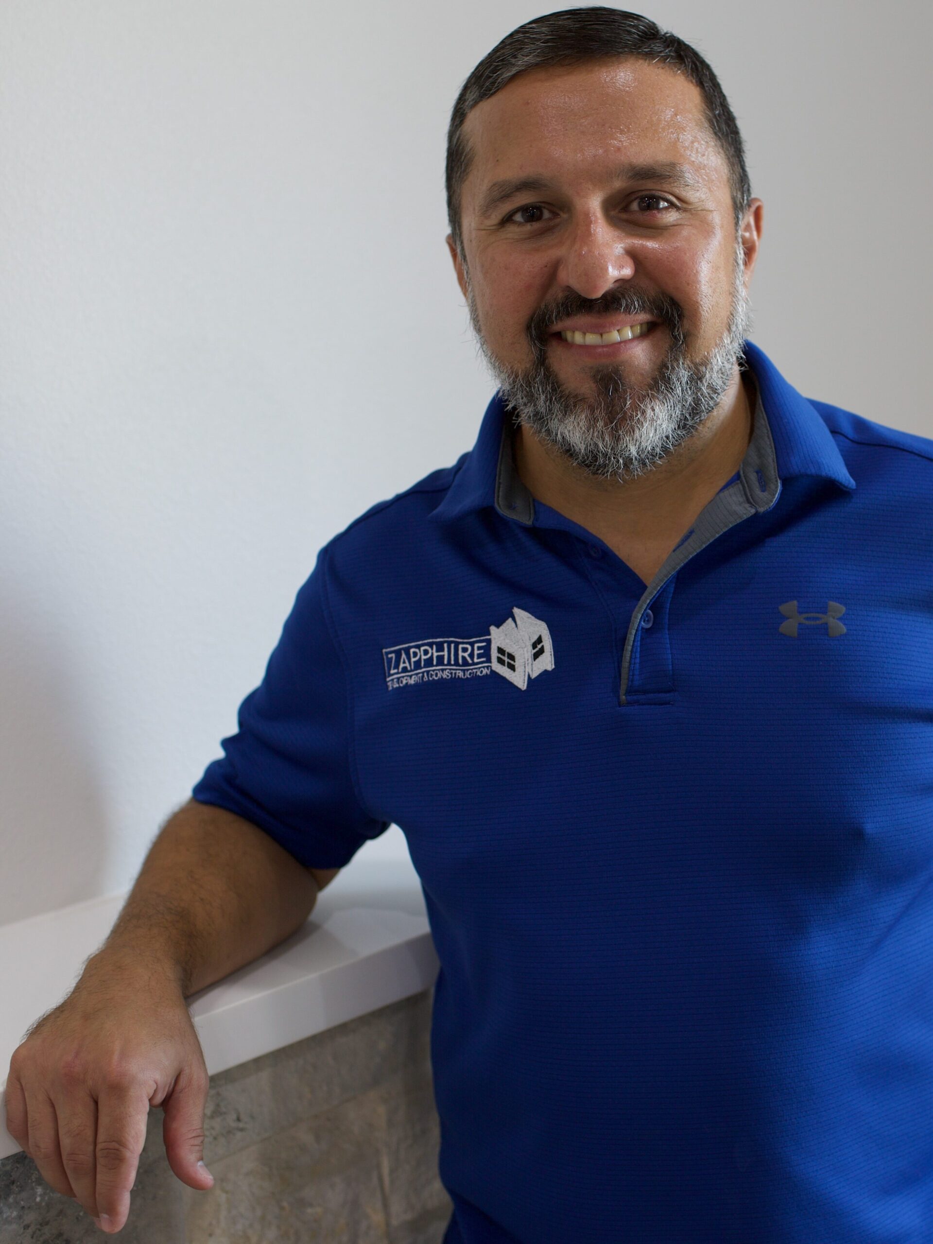 A smiling man with a grey beard is wearing a blue shirt in front of a grey wall.