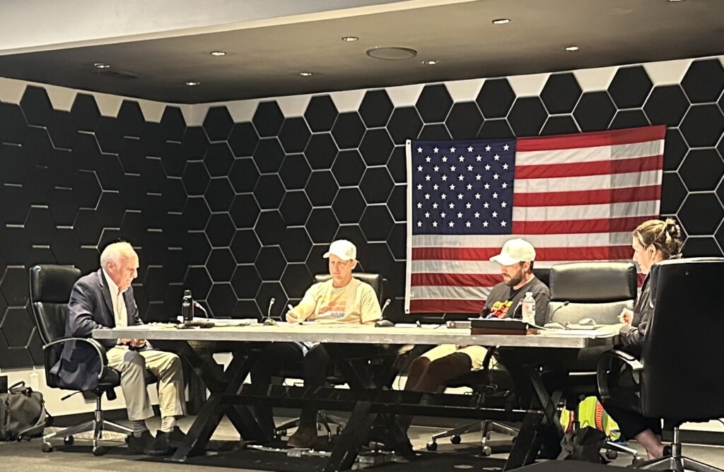 Four Starbase council members seated at a table during a public meeting with microphones and an American flag in the background.