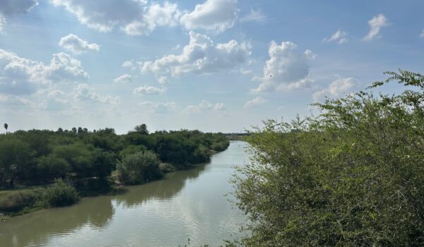 The Rio Grande River flowing between Texas and Tamaulipas near Nuevo Progreso, with dense green brush on both banks and scattered clouds overhead, illustrating the shared water source that sustains communities in the U.S. and Mexico.