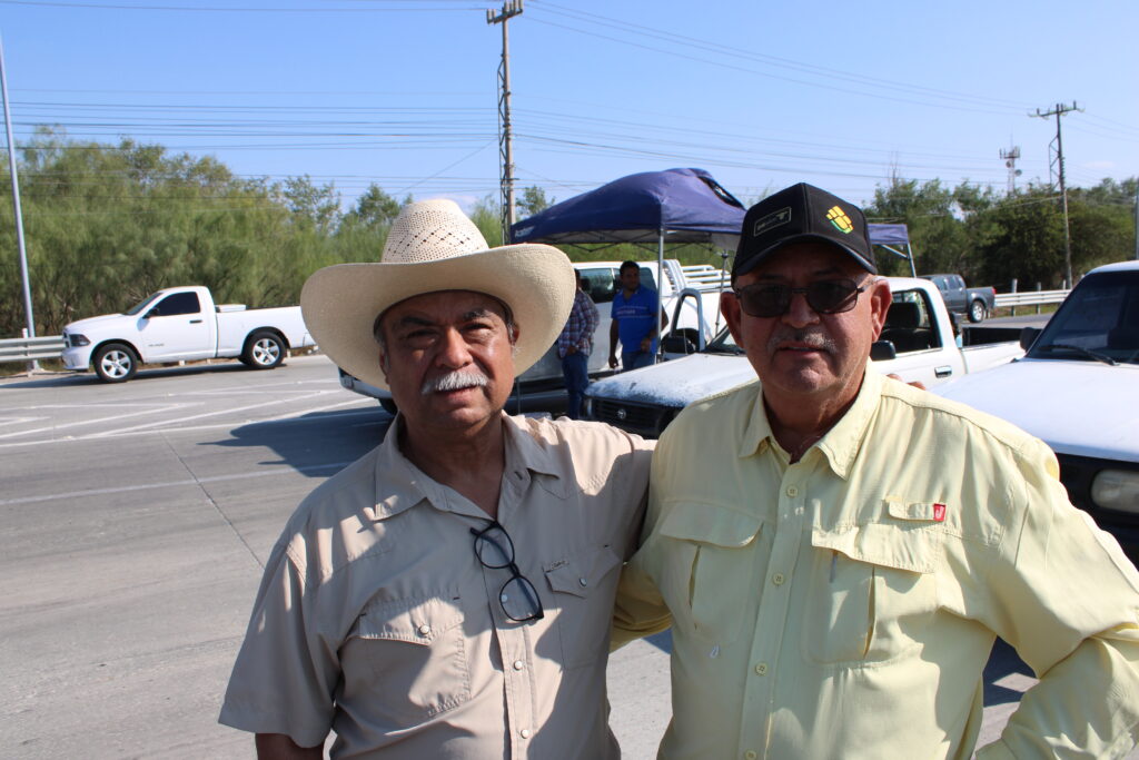 Two farmers standing together at the Reynosa-Pharr bridge access point during a protest blockade, with pickup trucks in the background.