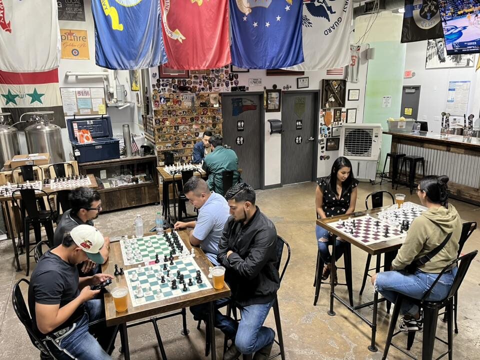 People play chess inside 5x5 Brewing Company’s taproom in Mission, Texas, with military flags hanging overhead.