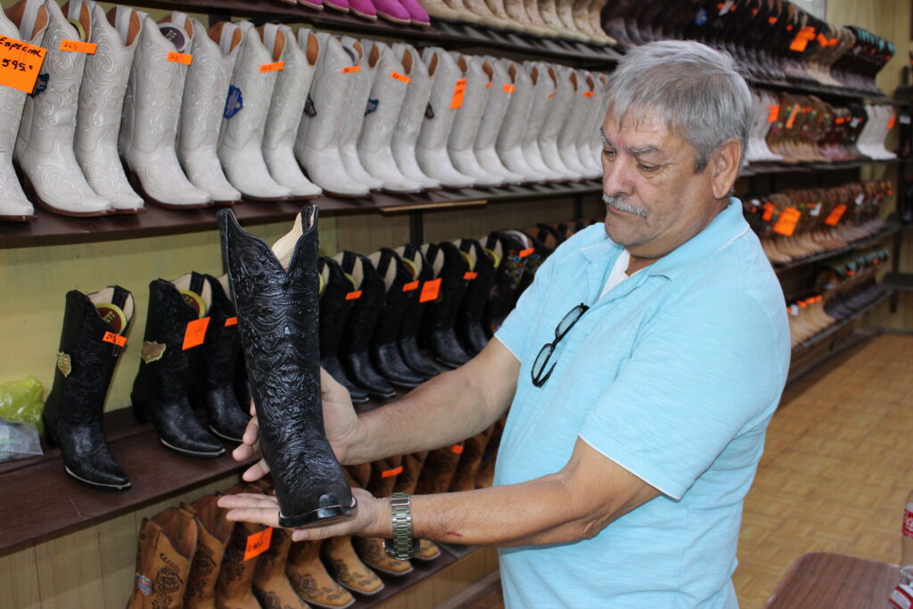 A bootmaker in Reynosa holds a black handcrafted boot while standing inside a shop lined with merchandise.