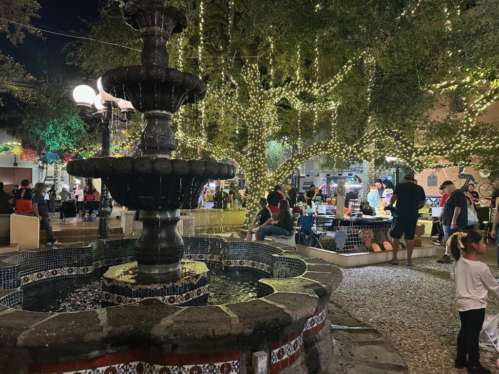 A large crowd gathers at Market Square in downtown Brownsville to watch performers during a First Friday celebration, surrounded by trees wrapped in string lights.