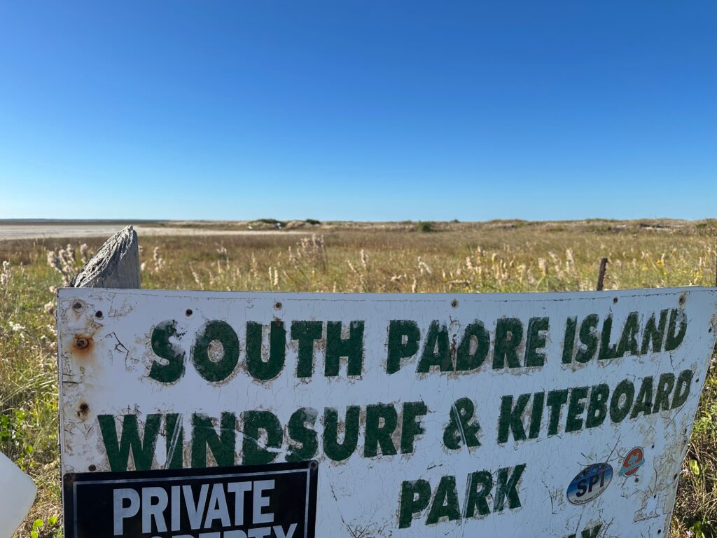 A weathered sign for the South Padre Island Windsurf & Kiteboard Park stands near dunes and grassland where TxDOT has proposed one possible causeway route.