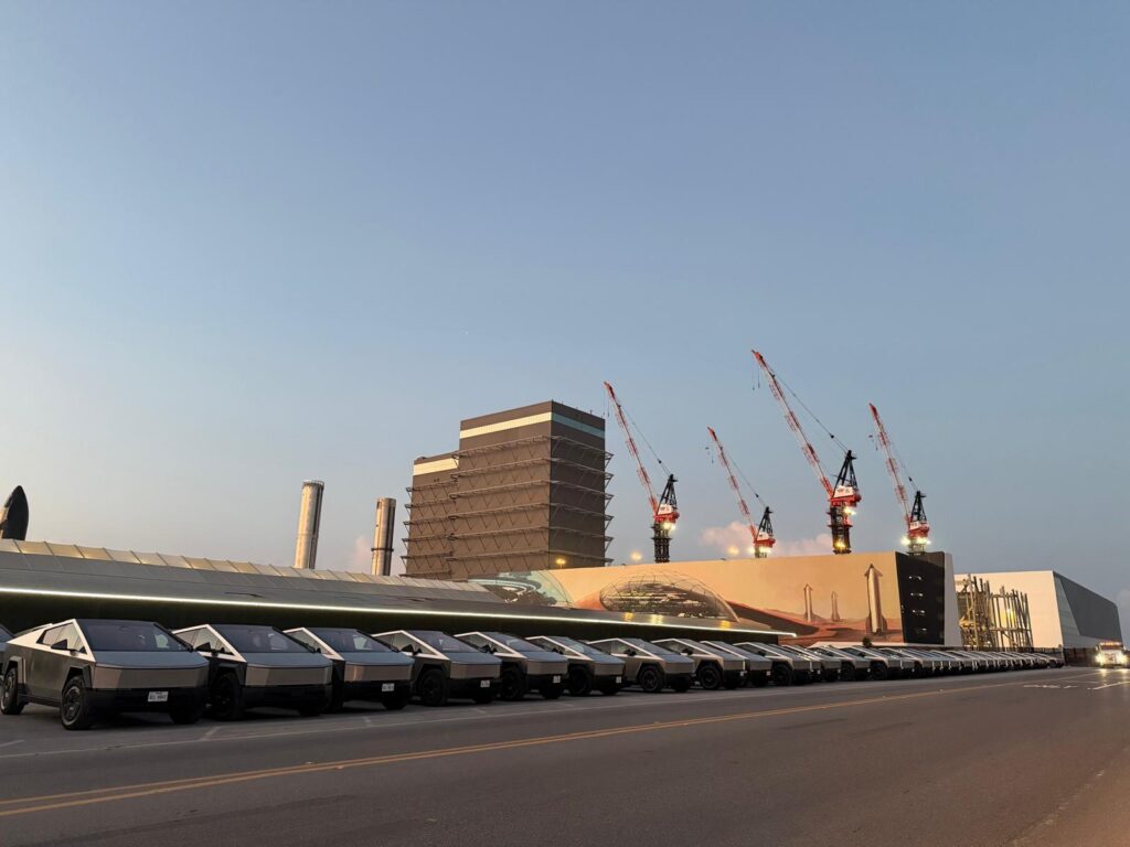 Multiple Cybertrucks parked in front of SpaceX buildings with cranes in the background.