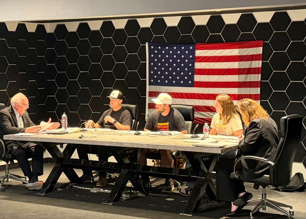 Five city officials seated at a table during a meeting, with an American flag displayed behind them.