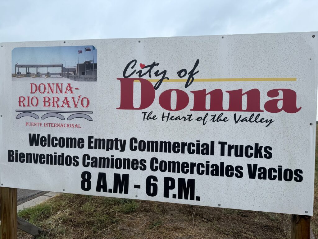 White roadside sign reading “Welcome Empty Commercial Trucks” with City of Donna branding near the international bridge.