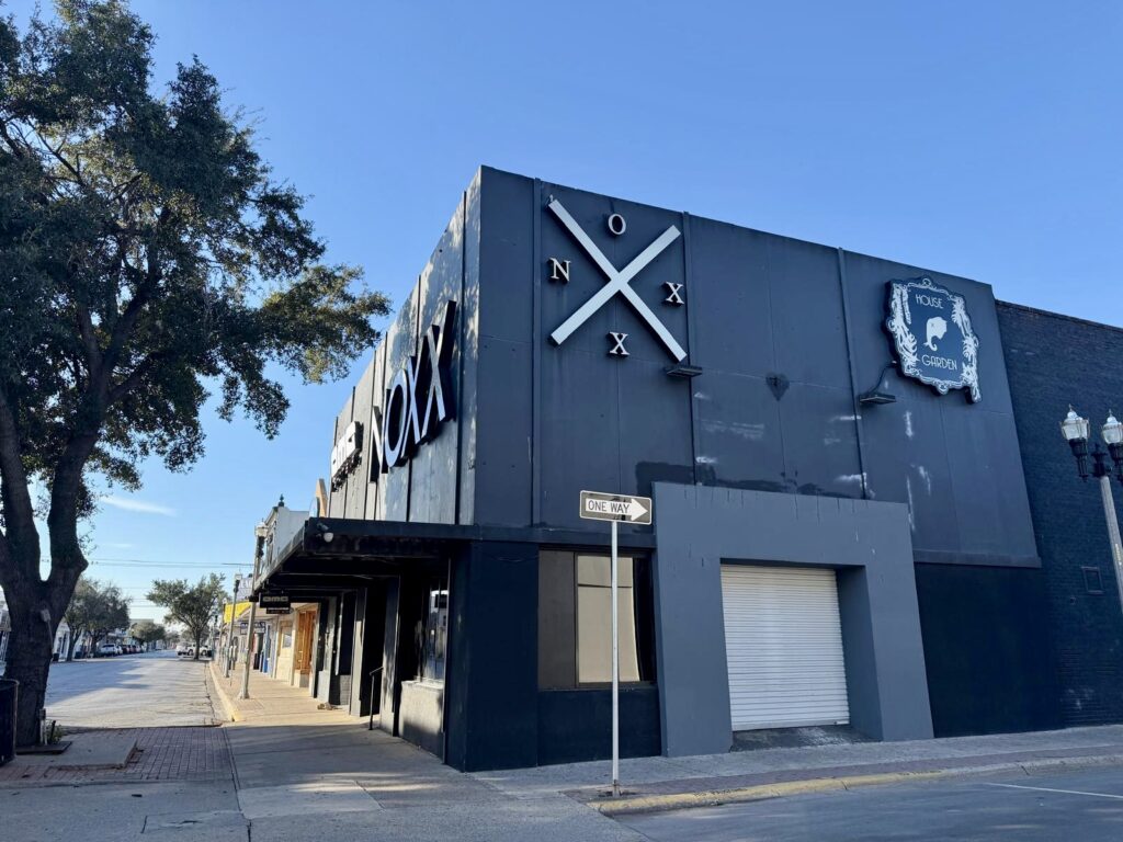 A dark gray building with large “NOXX” lettering on the facade along a downtown street in daylight.
