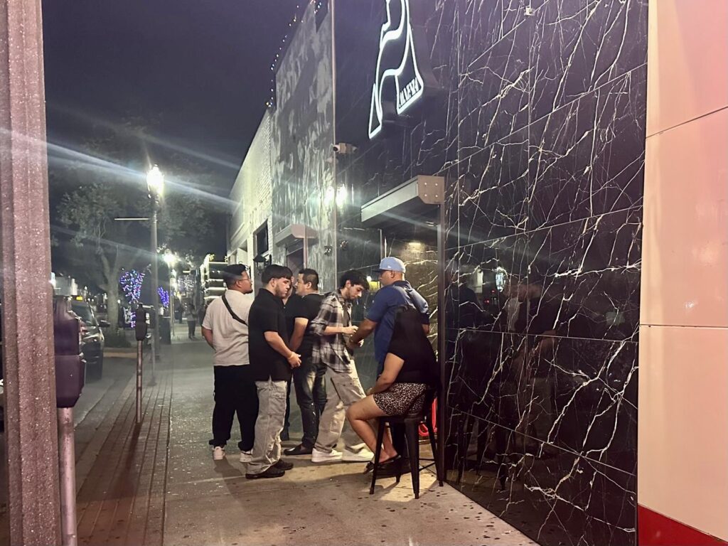 Several people standing and talking outside a nightclub entrance at night on a downtown sidewalk.