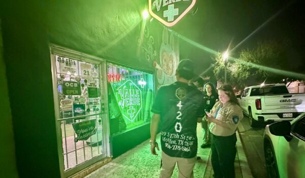 A woman in a khaki uniform speaking with a man outside a storefront with a bright green “Valle Verde” sign at night.