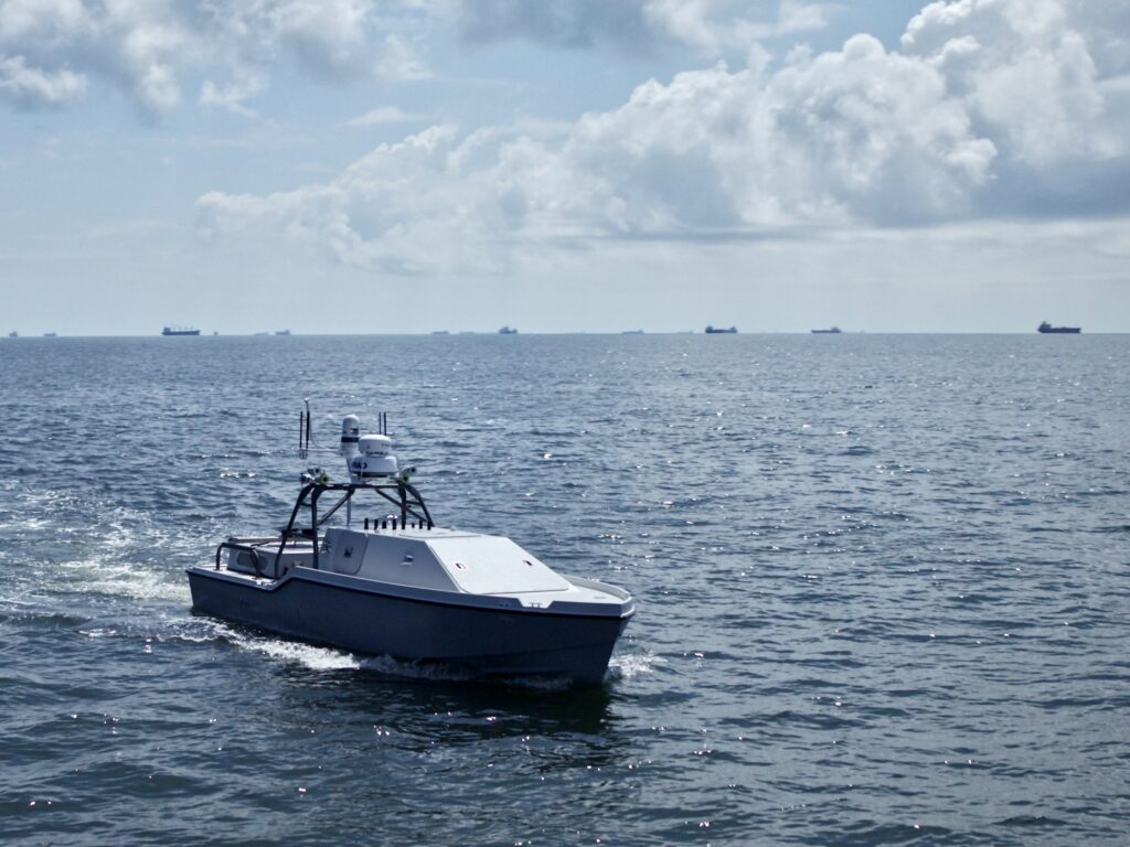 A gray unmanned boat moves through open water with large cargo ships visible on the distant horizon.