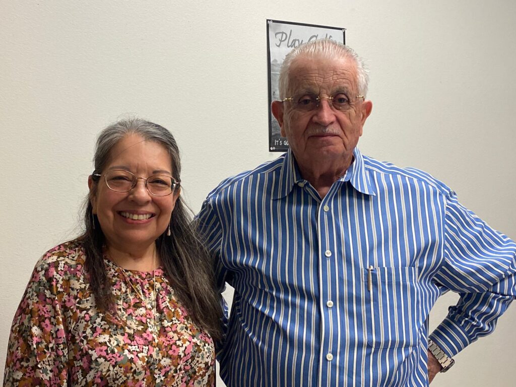 Irma Rosales and Carlos Martínez stand side by side indoors, smiling for a portrait photo.