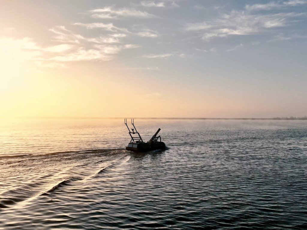 A robotic boat travels across calm water at sunset, silhouetted against an orange sky.
