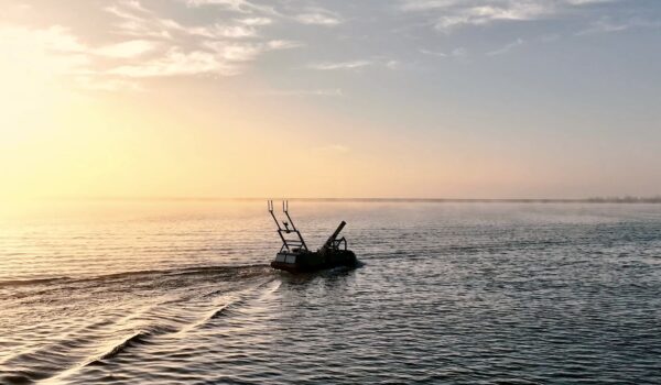 A robotic boat travels across calm water at sunset, silhouetted against an orange sky.