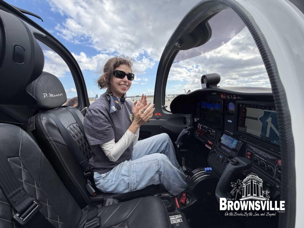 A young woman wearing sunglasses sits in the cockpit of a small training airplane, smiling and holding her hands up, with digital flight instruments and controls visible in front of her.