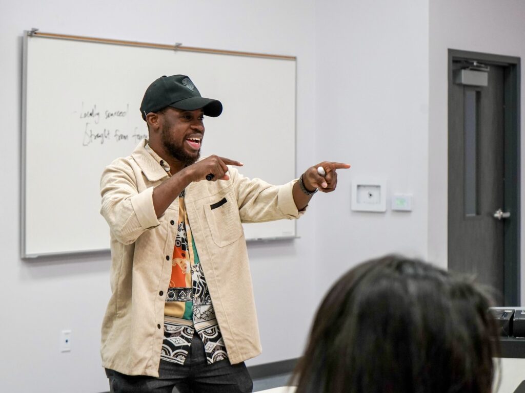 A man wearing a cap and beige jacket speaks animatedly while pointing toward participants during a small business workshop inside a classroom at the Center for Education and Economic Development in Mission.