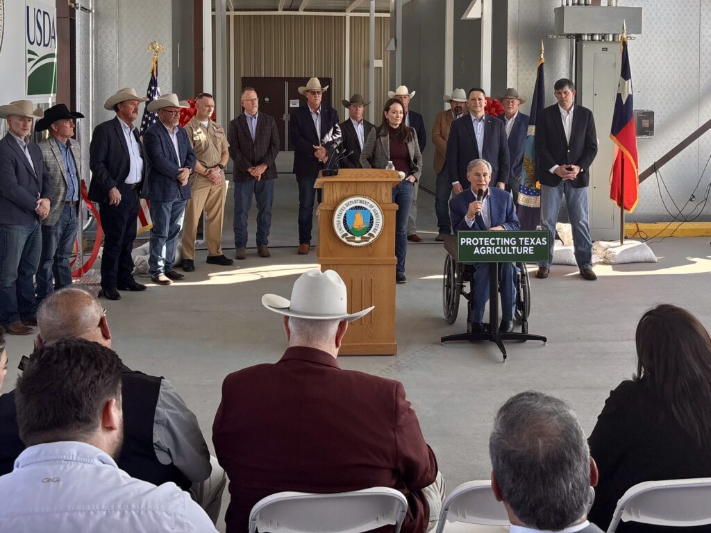 Texas Gov. Greg Abbott speaks into a microphone in front of state and federal officials and an audience inside a USDA facility at Moore Air Field.