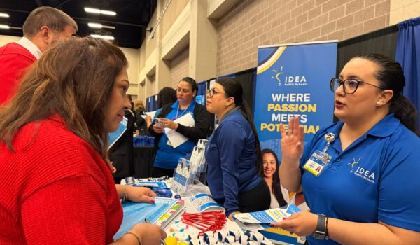 An IDEA Public Schools recruiter in a blue shirt talks with a woman holding brochures at a career fair booth with promotional materials and giveaways.