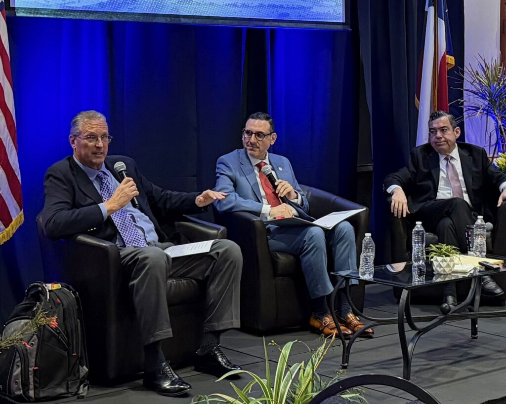 Tres ponentes están sentados en un escenario con micrófonos en la mano durante un debate sobre comercio, con iluminación azul y banderas colocadas detrás de ellos.