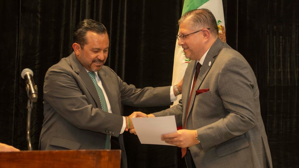 Jesús Salvador González Martínez presents recognition to Mission Economic Development Corp. CEO Teclo Garcia during a ceremony in front of a Mexican flag backdrop.