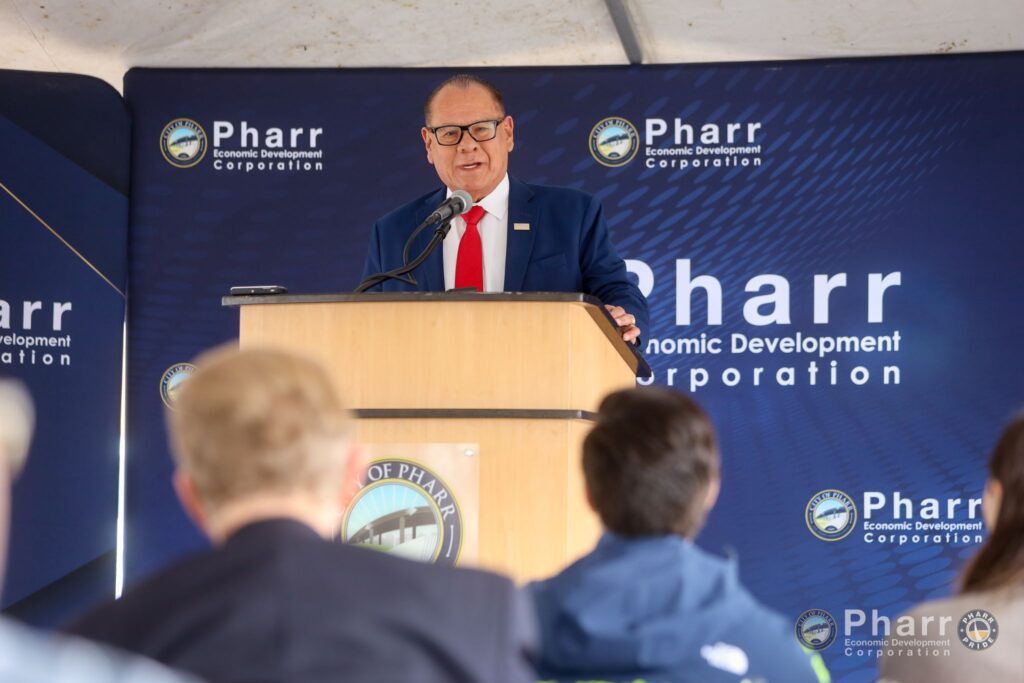 A man in a suit stands at a podium speaking to an audience, with Pharr Economic Development Corporation logos displayed behind him.