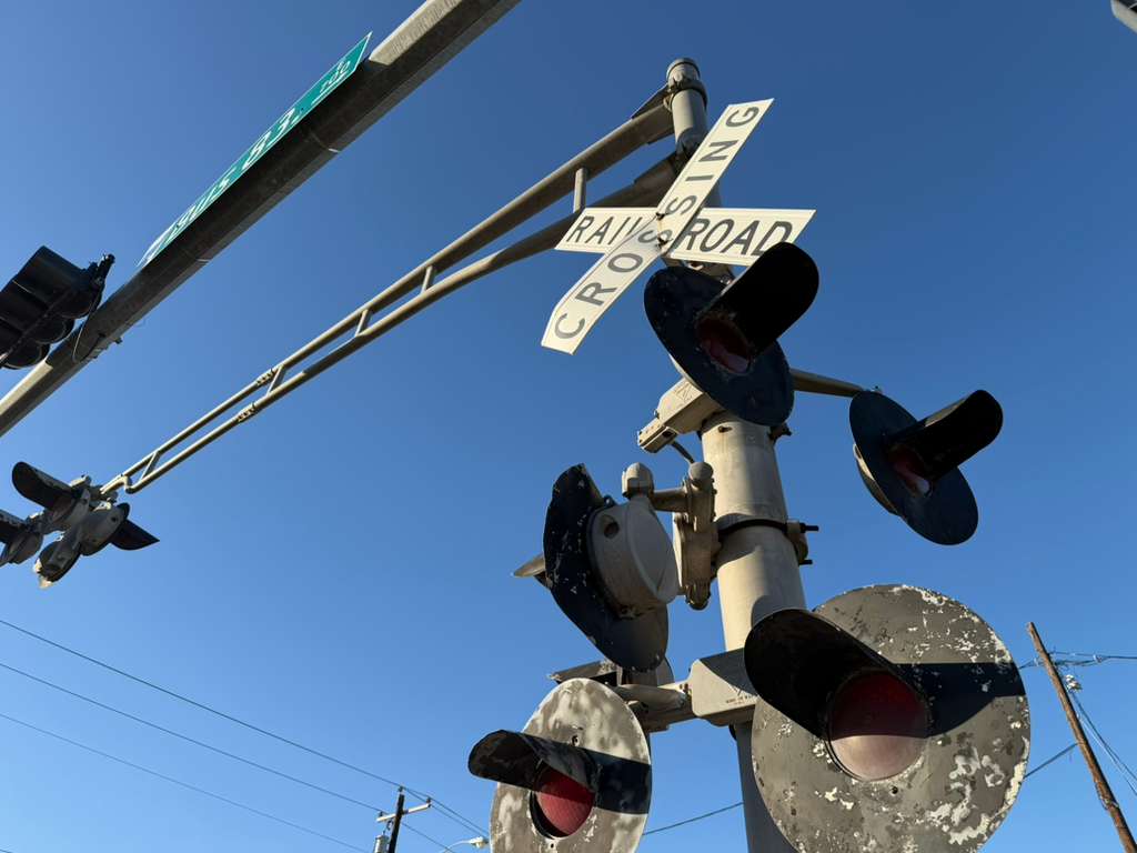A railroad crossing signal with warning lights and crossbuck signage is seen against a clear blue sky along a rail line in the Rio Grande Valley.