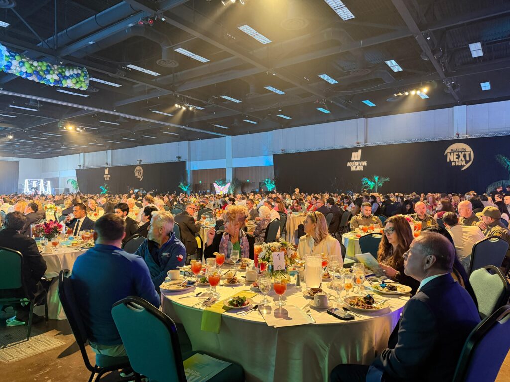 Hundreds of attendees sit at decorated banquet tables inside the McAllen Convention Center during the State of the City event, with stage lighting and large backdrop displays visible across the hall.
