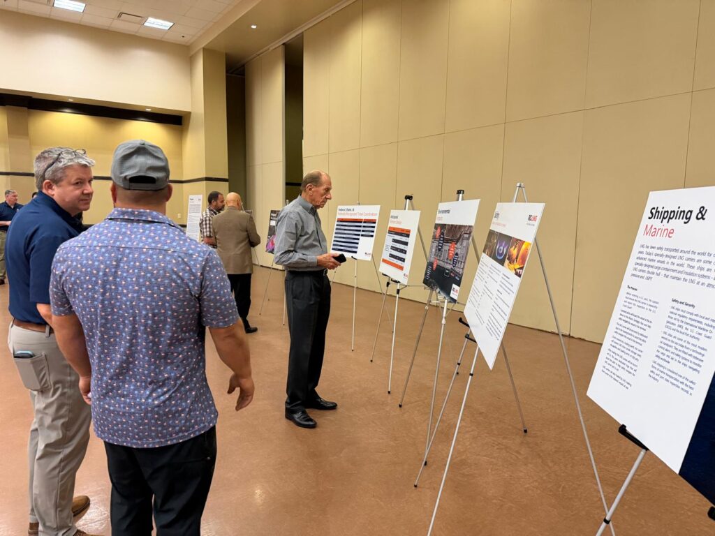 Several men stand in a conference room reading large presentation boards displayed on easels during a public meeting about the Rio Grande LNG project expansion.