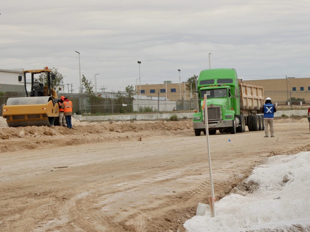 A dump truck, road roller, and construction workers in safety vests operate on a dirt construction site with a large industrial building in the background.