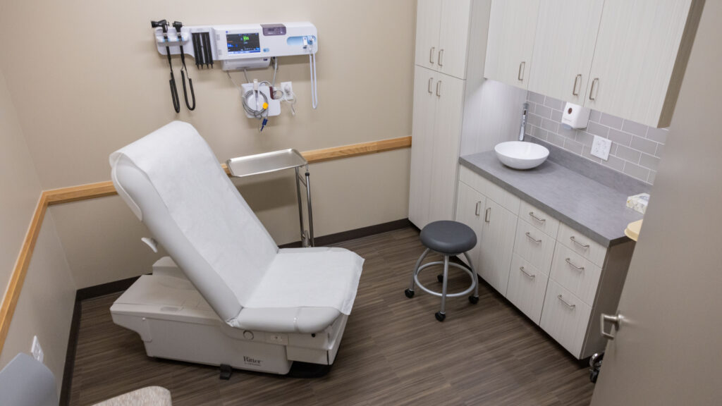 Interior of a medical exam room with an exam chair, mounted diagnostic tools, cabinets and a sink inside an H-E-B Wellness Primary Care clinic.