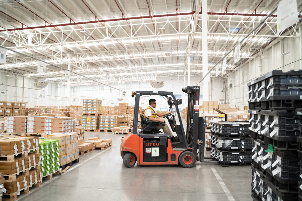 A worker drives a forklift through a large warehouse filled with stacked boxes and industrial shelving at a Valeo facility.