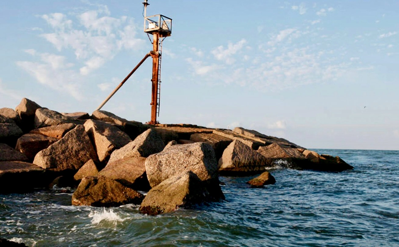 Rock jetty at Port Mansfield with waves hitting the shoreline and a navigation beacon structure above the rocks, captured from a surf forecast camera under a partly cloudy sky.