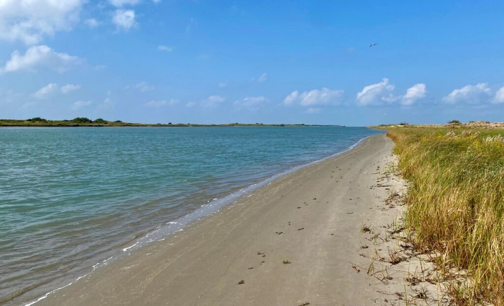 View looking west along the Mansfield Channel, showing calm blue water bordered by a narrow sandy shoreline and coastal grasses under a partly cloudy sky.