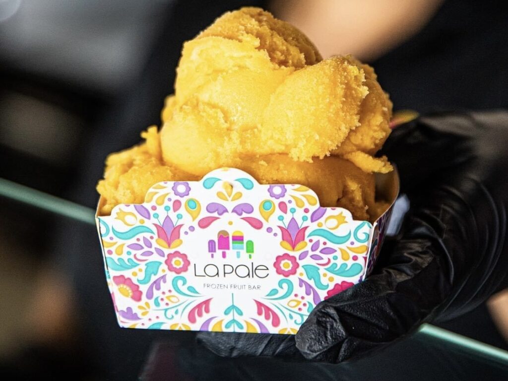 Scoops of orange-colored ice cream served in a colorful La Pale Frozen Fruit Bar container held by a gloved hand.