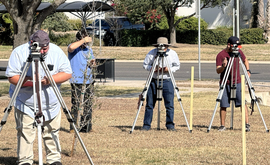 Several students stand behind tripod-mounted surveying instruments in a grassy area while recording measurements during a training exercise.