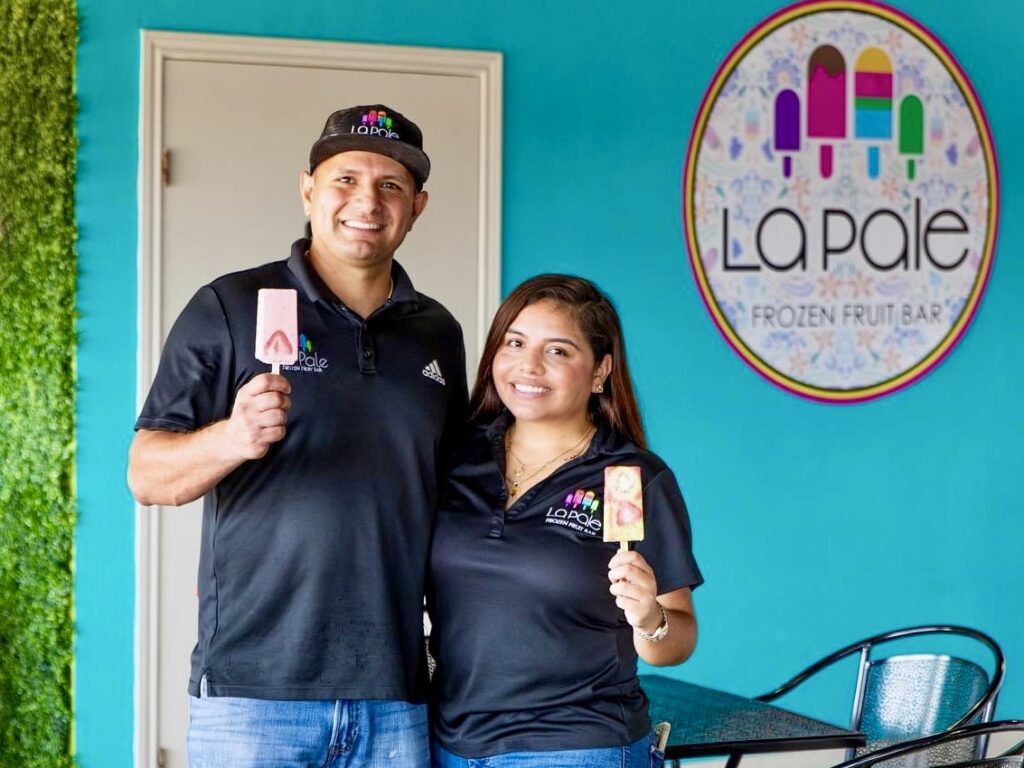Gerardo and Daisy Alcazar stand inside their Brownsville paletería holding fruit paletas in front of a La Pale Frozen Fruit Bar sign.