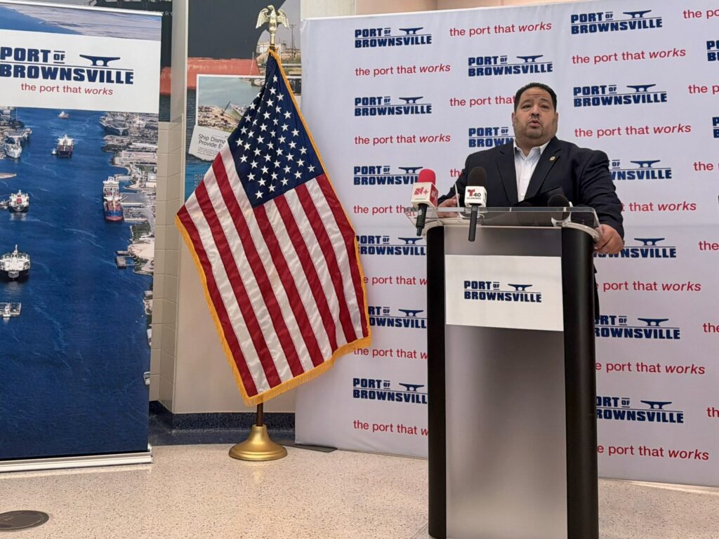 Port of Brownsville Chairman Steve Guerra speaks at a podium during a media briefing with a U.S. flag and Port of Brownsville backdrop behind him.