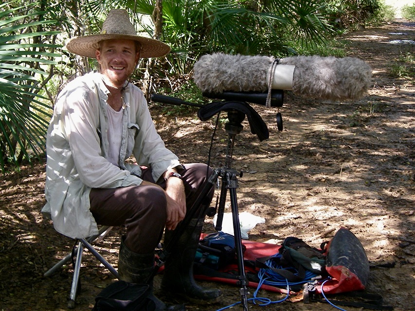 A researcher wearing a wide-brimmed hat sits beside audio recording equipment in a wooded field site, surrounded by vegetation, as he studies bird sounds in Venezuela.