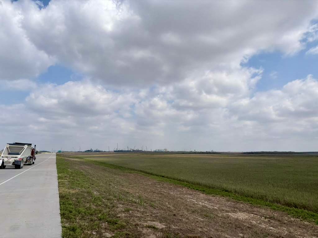 Road and coastal marshland near the Port of Brownsville with industrial facilities visible in the distance.