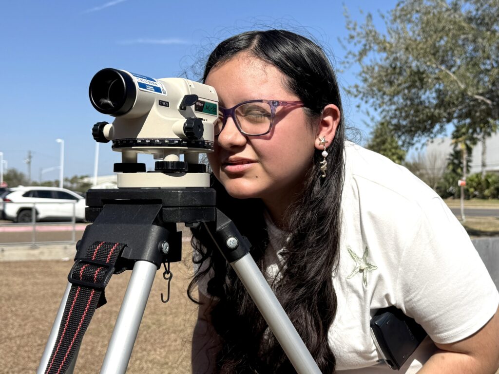 A student peers through a surveying instrument mounted on a tripod while practicing field measurement techniques outdoors.