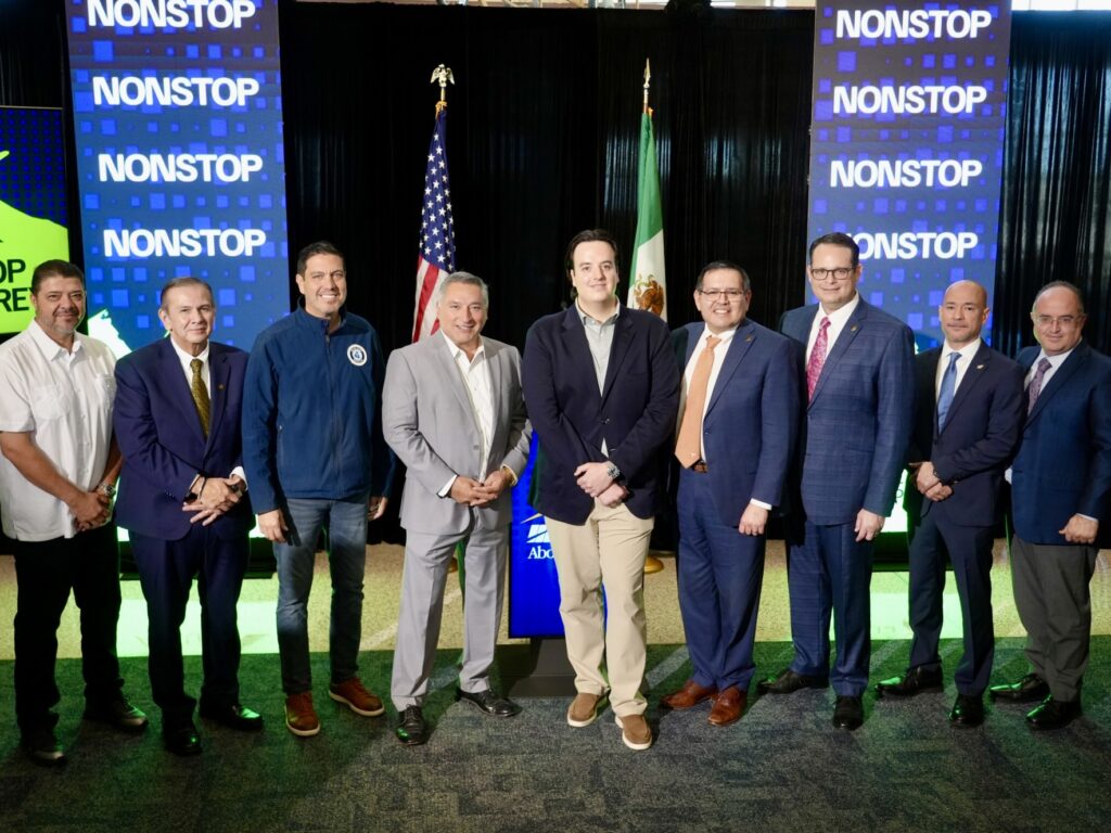 Group of local officials and airline representatives stand together on stage at McAllen International Airport during the announcement of a new nonstop flight between McAllen and Monterrey, with U.S. and Mexican flags behind them.