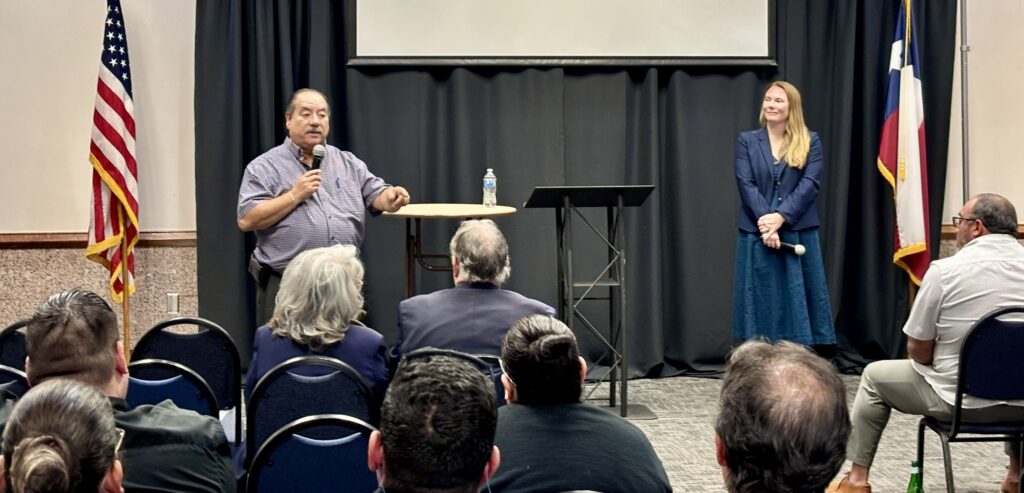 Tony Aguirre habla por un micrófono mientras se dirige a un público sentado en una sala de reuniones.